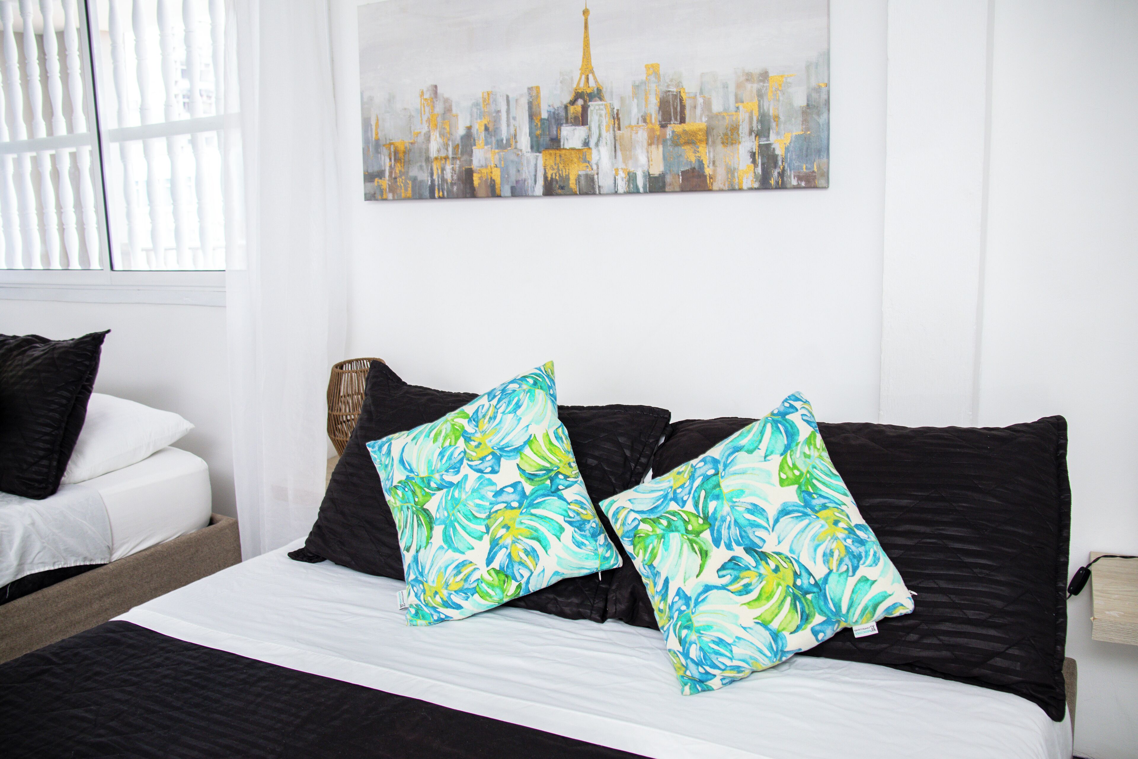 A neatly made bed with black and white bedding, two colorful pillows, and a cityscape painting on the wall.