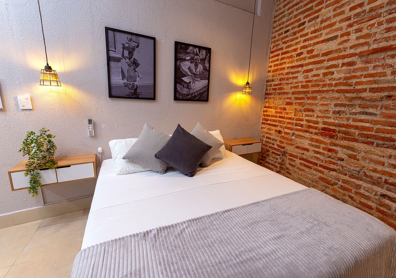 A bedroom with a bed, pillows, a bedside table, a wall-mounted light, and a brick wall.
