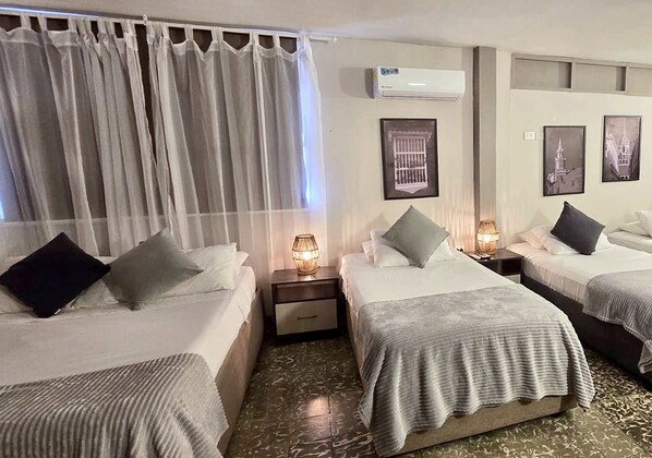 A hotel room with two beds, a nightstand with a lamp, a window with curtains, and an air conditioning unit on the wall.