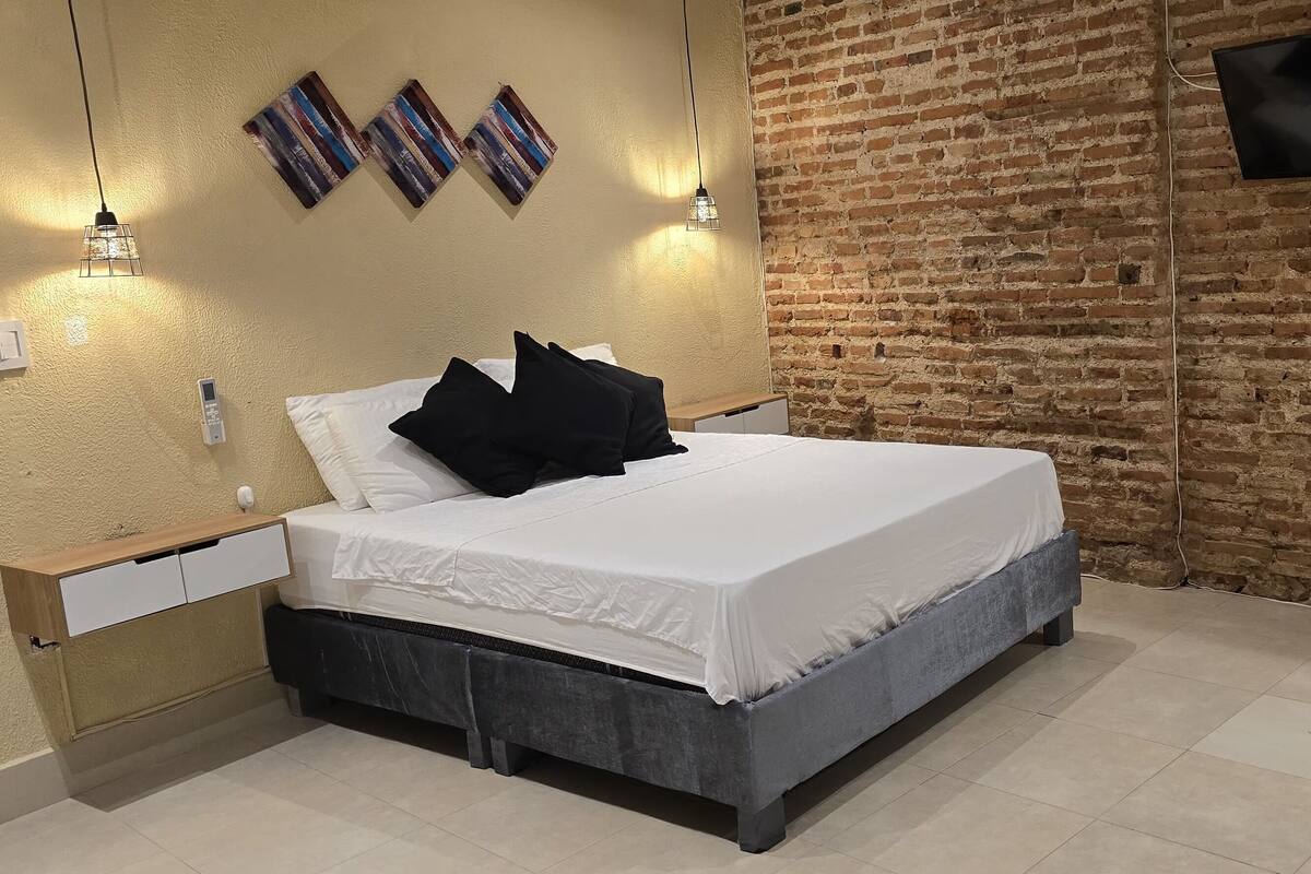 A modern bedroom with a brick wall, a bed with white bedding, black pillows, a bedside table, a TV, and a desk with a chair.