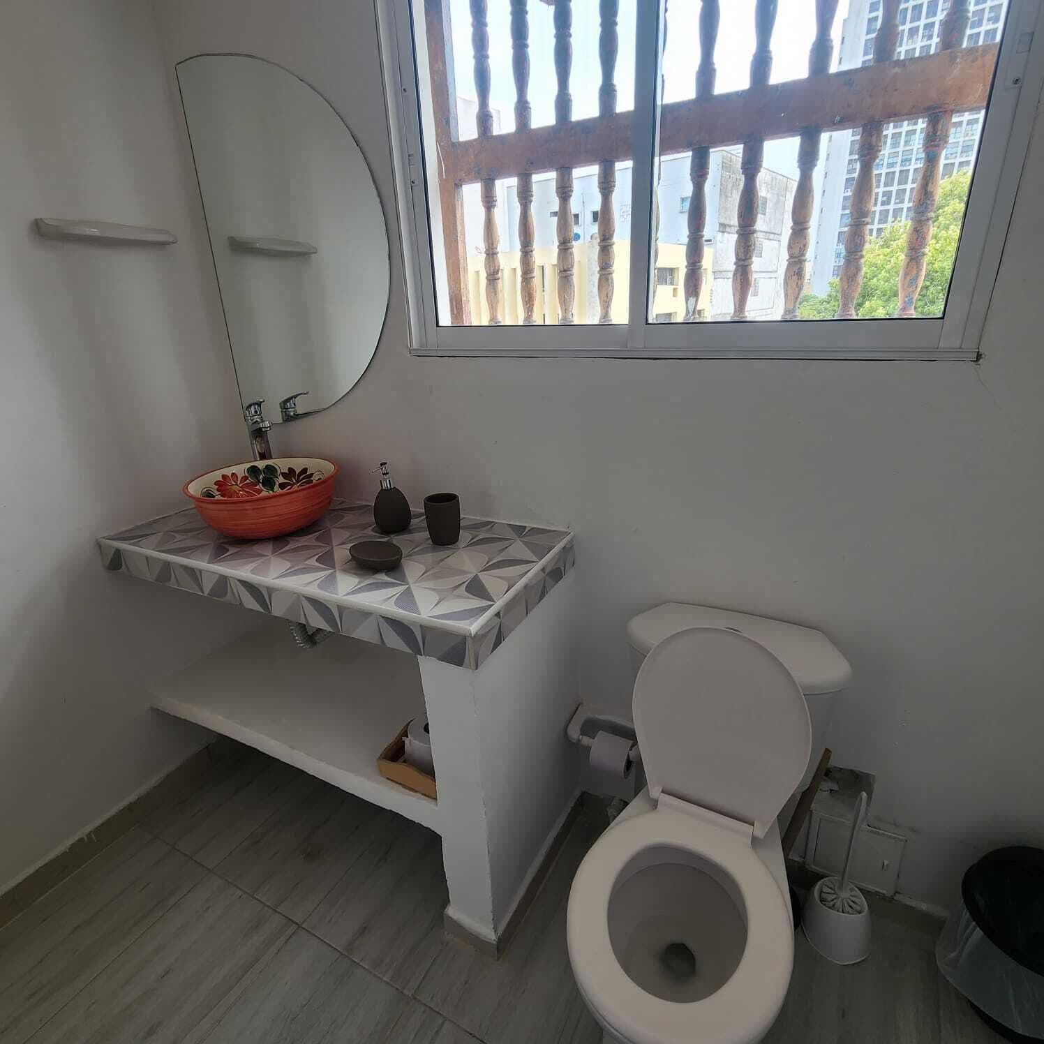 Ein Badezimmer mit einer weißen Toilette, einem runden Spiegel, einem roten Waschbecken und einem Fenster mit Blick auf Gebäude.