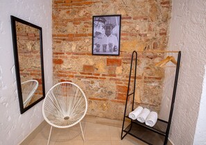 A room with a brick wall, a white chair, a mirror, a framed photo, and a towel rack.