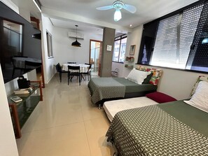 A room with two beds, a dining table with chairs, a ceiling fan, and a window with blinds.