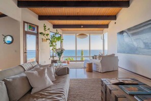 A spacious living room with a large window overlooking the sea, a modern sofa, and a wooden ceiling. - A Beachfront Bungalow on Malibu's Broad Beach (Malibu)