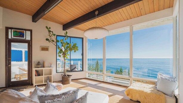 Ein Zimmer mit großem Fenster zum Meer, ein weißes Sofa mit gestreiften Kissen, Holzboden und einer hängenden Lampe.