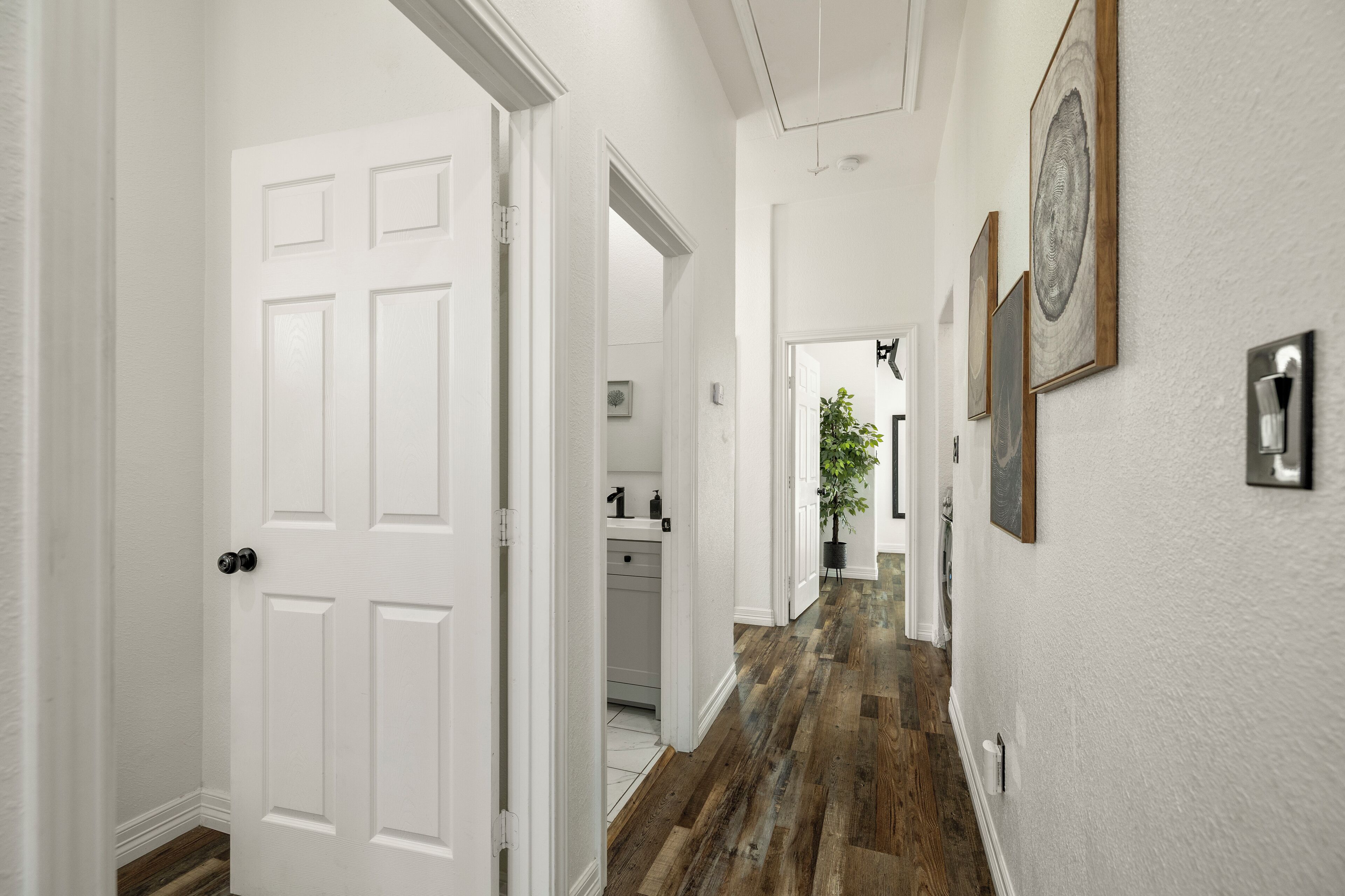 Un couloir aux murs blancs, doté d’un plancher en bois et d’une porte blanche. Des tableaux encadrés ornent le mur, et une plante est visible au loin.