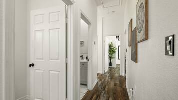Un couloir aux murs blancs, doté d’un plancher en bois et d’une porte blanche. Des tableaux encadrés ornent le mur, et une plante est visible au loin.