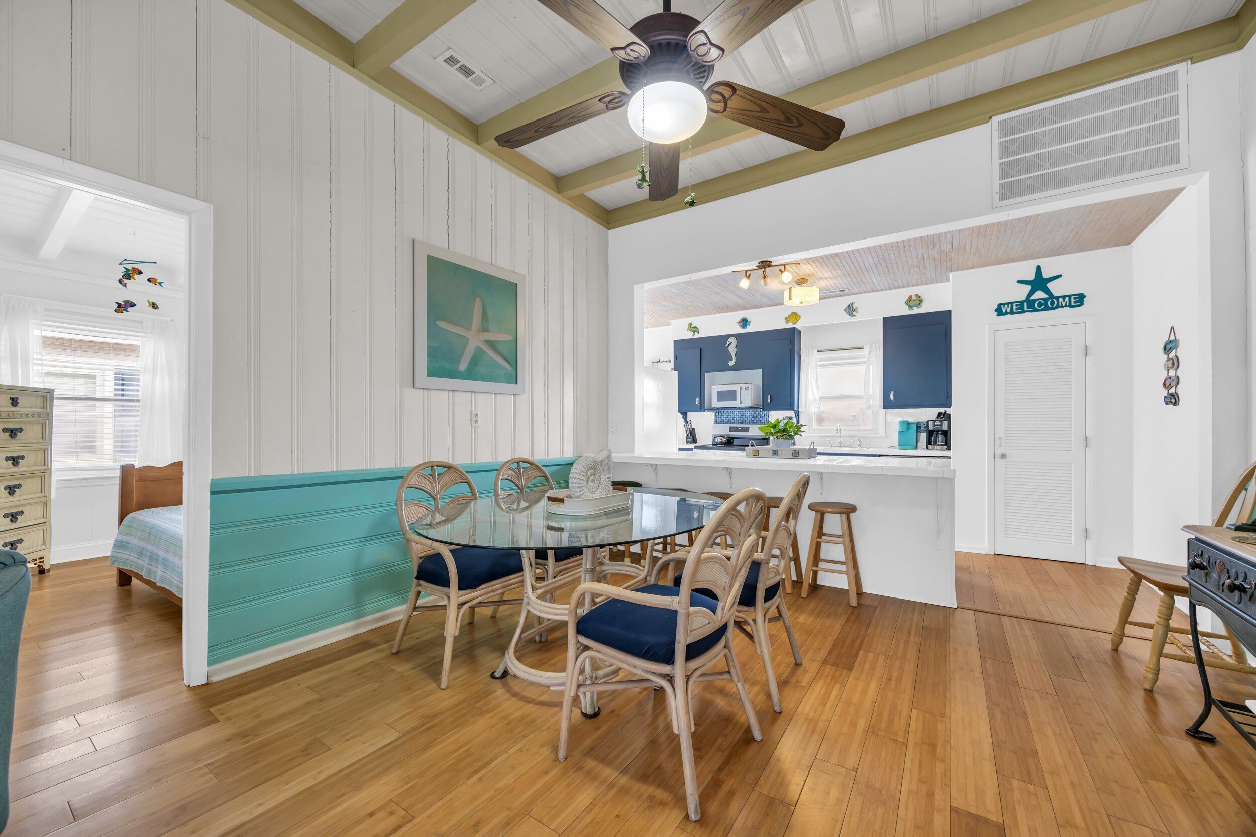 A cozy room with a glass dining table, wicker chairs, a teal bench, wooden floors, a ceiling fan, and a kitchen area with a microwave and a coffee maker.