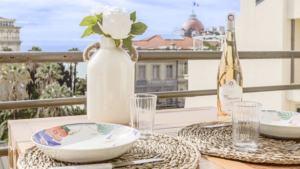 A balcony with a table set for two, featuring a white vase with a white flower, a bottle of champagne, and two glasses. The background includes a view of a building with a dome and a flag. - Le Mirazur by Interhome (Nice)