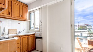 A compact kitchen with wooden cabinets, a white tiled backsplash, a window, and a view of the city through the balcony. - Le Mirazur by Interhome (Nice)