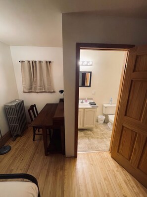 Una habitación con una mesa de comedor de madera, sillas, un radiador y una puerta de madera que da a un baño con inodoro y lavamanos.