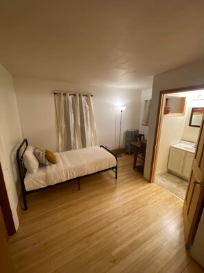 Un dormitorio con una cama individual, una mesita de noche, un radiador y un baño visible a través de una puerta abierta.