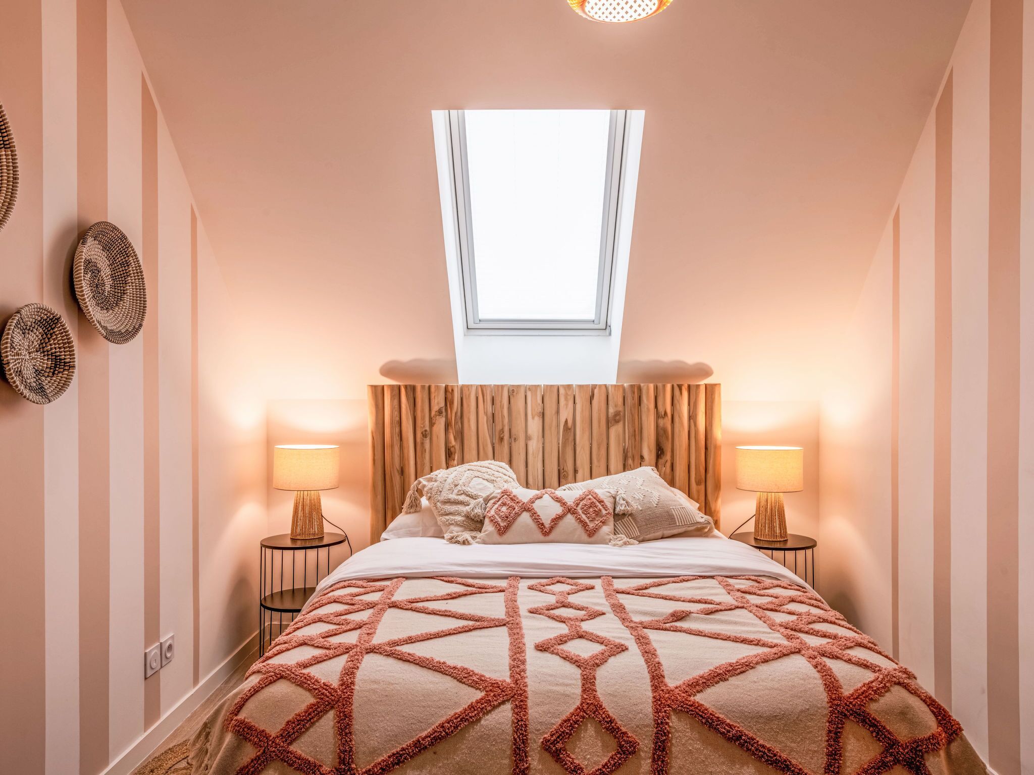 Een slaapkamer met een schuine kap, een dakraam, een bed met een bedspreider met een geometrisch patroon, twee bedlampen en een wandlamp.