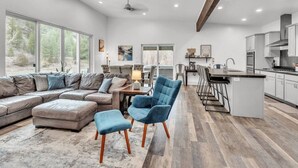 Amplia sala de estar con un sofá grande en forma de L, un sillón azul y un pequeño otoman. La habitación tiene piso de madera, un ventanal con vista a árboles, y una zona de cocina con armarios blancos y diseño moderno.