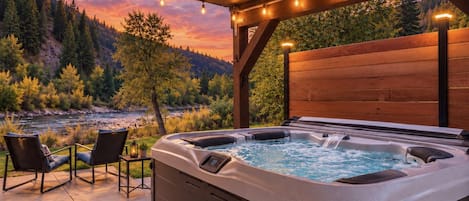 Un bain à remous doté d’un panneau de commande, de deux fauteuils et d’une terrasse en bois offrant une vue sur une rivière et une forêt.
