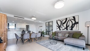 A modern living room with a grey sofa, a coffee table, and a dining area with chairs and a table. There are abstract paintings on the wall and a floor lamp.