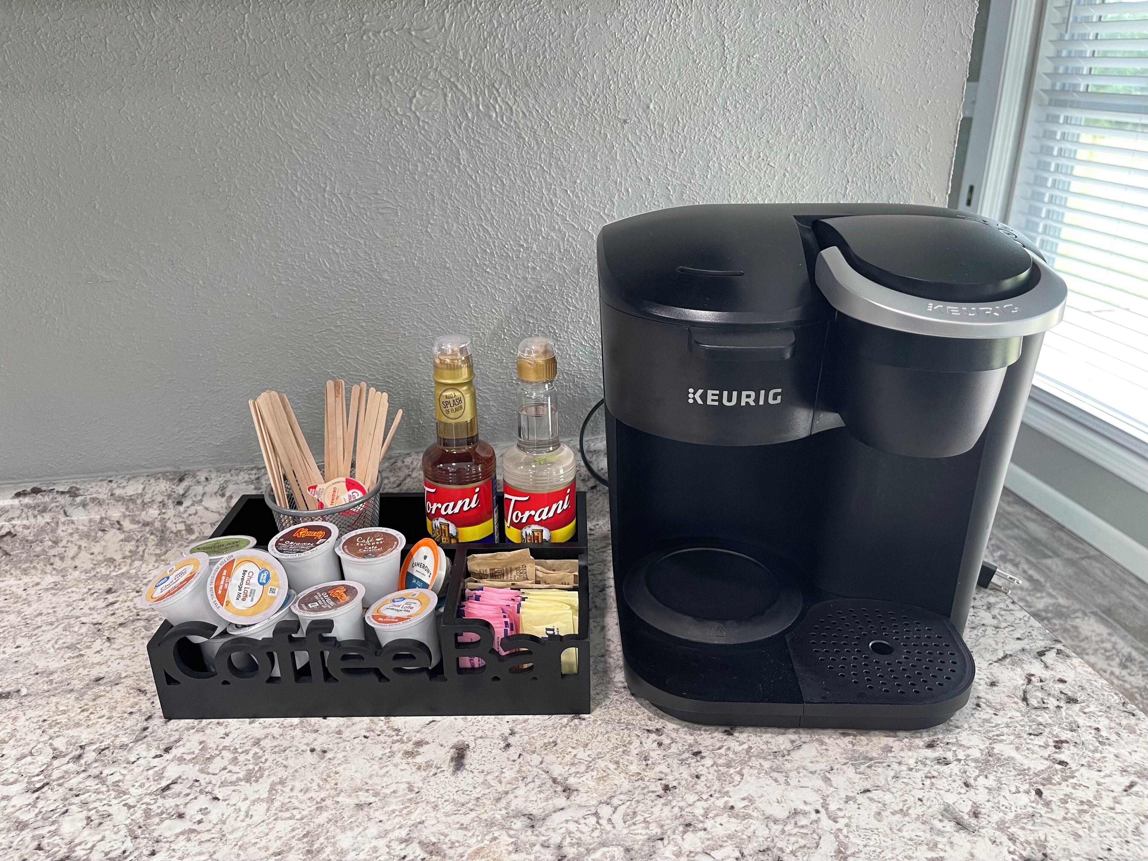 Una cafetera Keurig y una estación de café con varios tipos de cápsulas, siropes Torani y palitos de madera para remover sobre una encimera de granito.