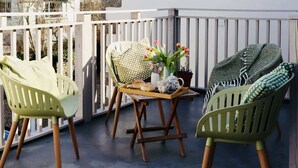 Un balcon avec deux chaises vertes, une table en bois sur laquelle se trouve un plateau de pâtisseries, un vase rempli de fleurs et une couverture jetée sur l’une des chaises.