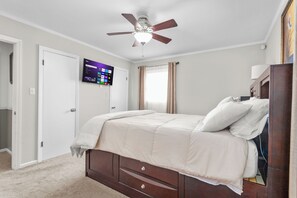 Ein Schlafzimmer mit einem Bett, einem Deckenventilator, einem an der Wand montierten Fernseher und einem Fenster mit Vorhängen.