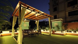 Un área de estar al aire libre con un pérgola de madera, asientos acolchados e iluminación ambiental.