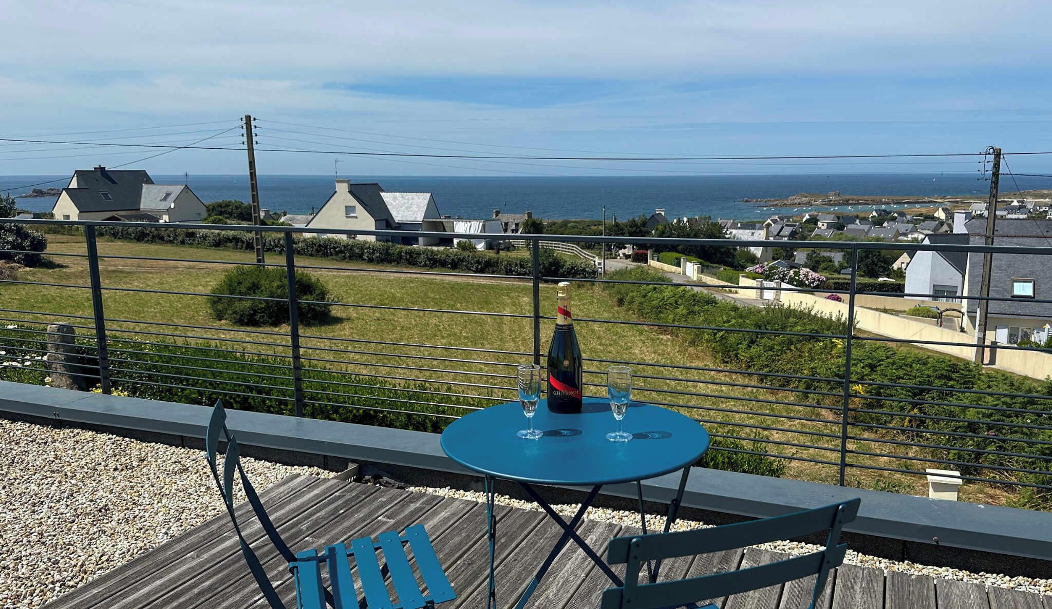 A balcony with a table, a bottle of wine, and a view of the sea and houses.