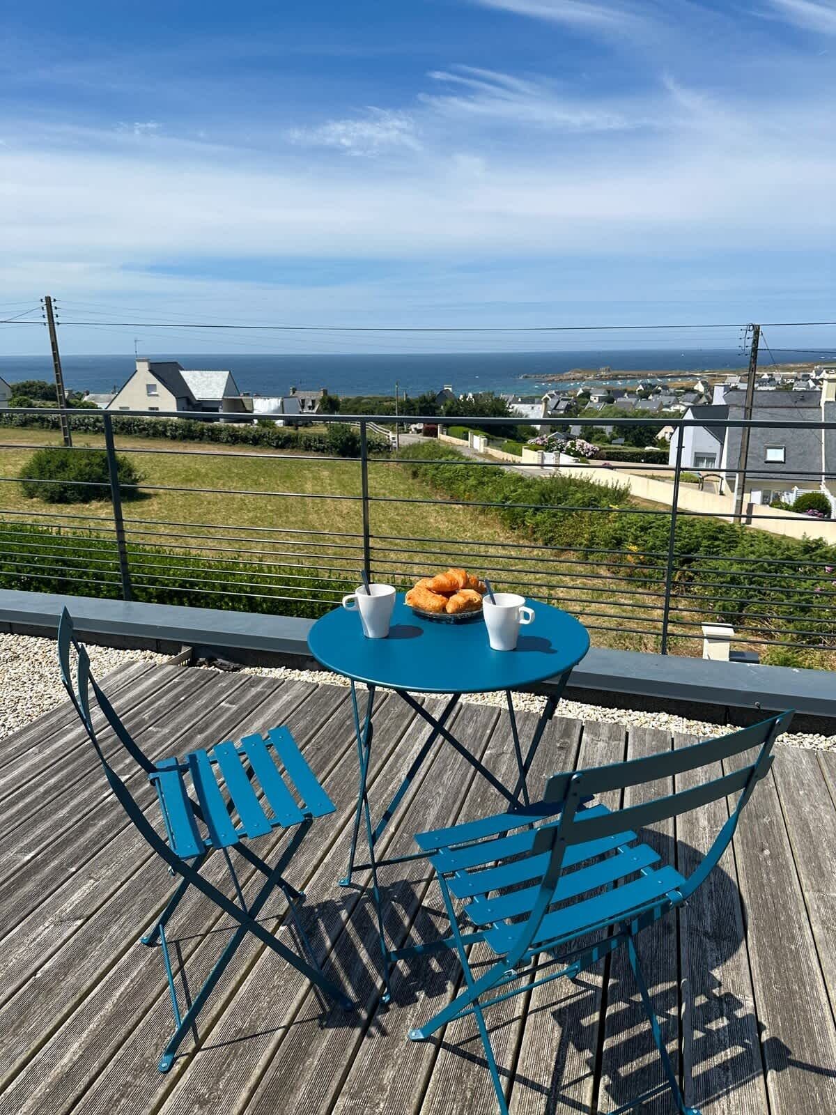 Et blåt metalbord og stole på en træterrasse med udsigt over en kystby med huse og havet.