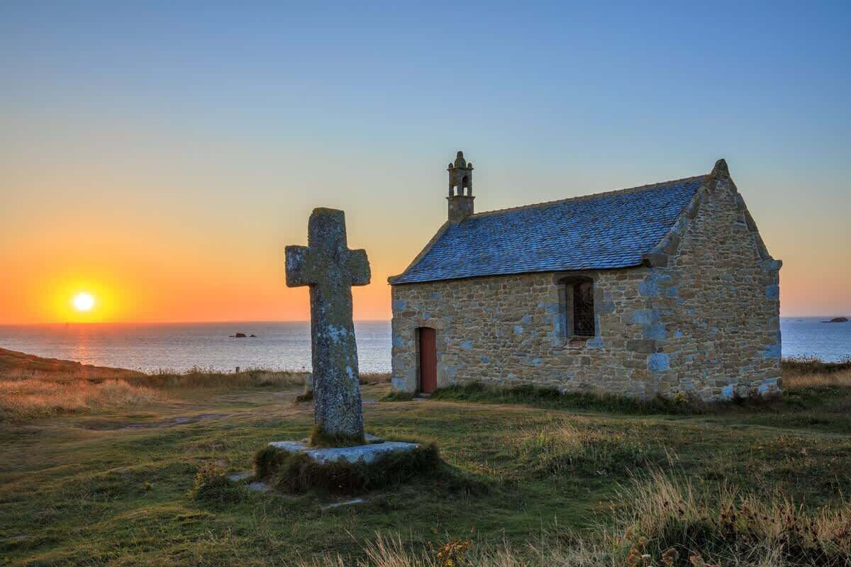 Eine steinerne Kirche mit Glockenturm und Kruzifix in einem grasbewachsenen Feld bei Sonnenuntergang.