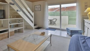 A living room with a TV, shelves, wicker baskets, a glass coffee table, and a sliding glass door leading to an outdoor patio with a table and chairs. - L'Échappée Chaumoise  Maison Cosy à 70m de la Plage (Les Sables-d´Olonne)