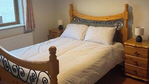 Una cama bien hecha con tablero de cabecera y pie de cama de madera, dos mesitas de noche con lámparas y una ventana con cortinas.
