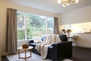 A modern living room with a sofa, a small table, and a view of a garden through large windows. - Skyline Haven: Green View, Bird Songs, Parking, Espresso & Netflix (Wellington)