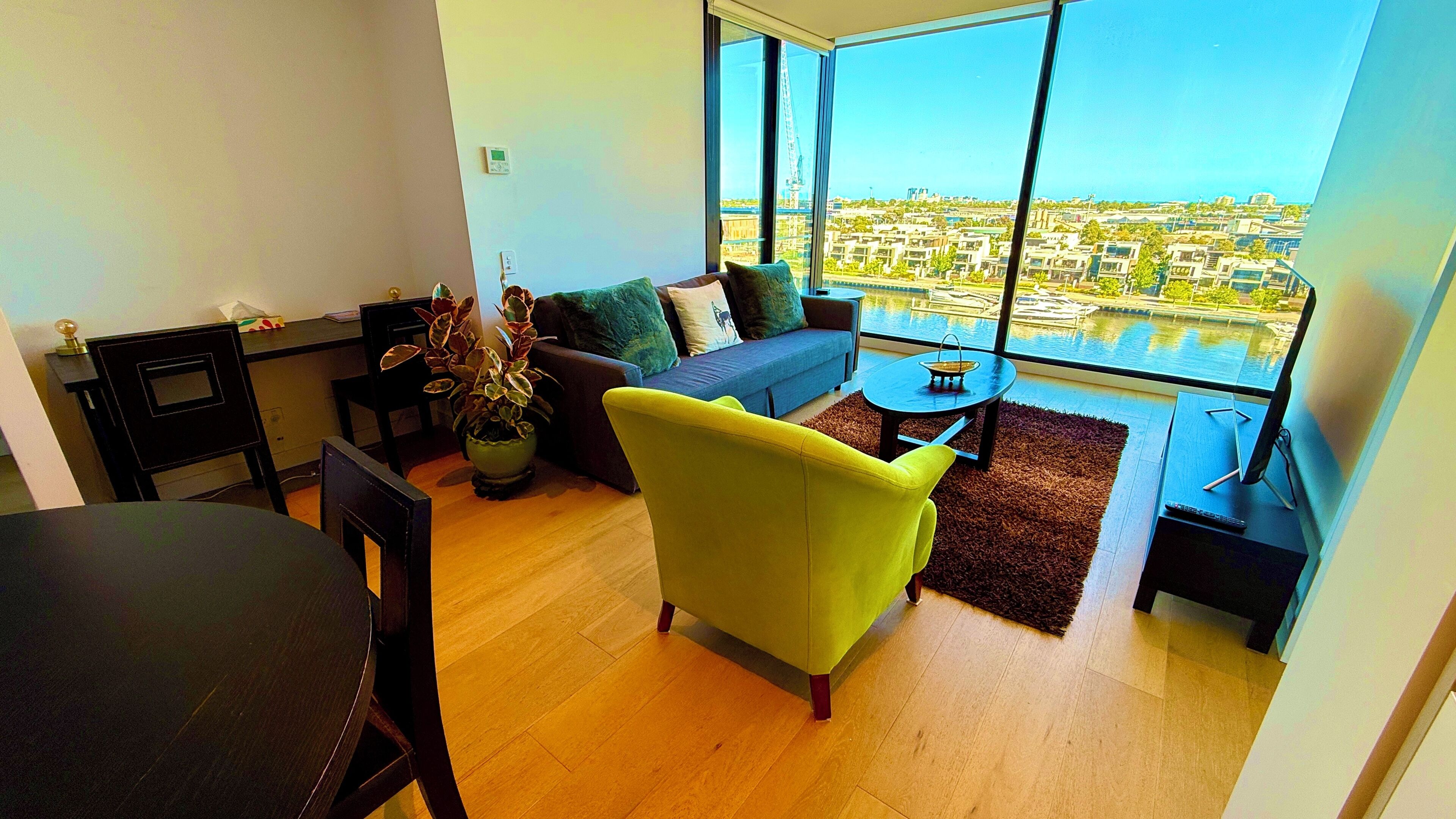 Una sala moderna con sofá, sillón y mesa de centro, un comedor con mesa y sillas, y un gran ventanal con vistas a un paisaje urbano y al agua.