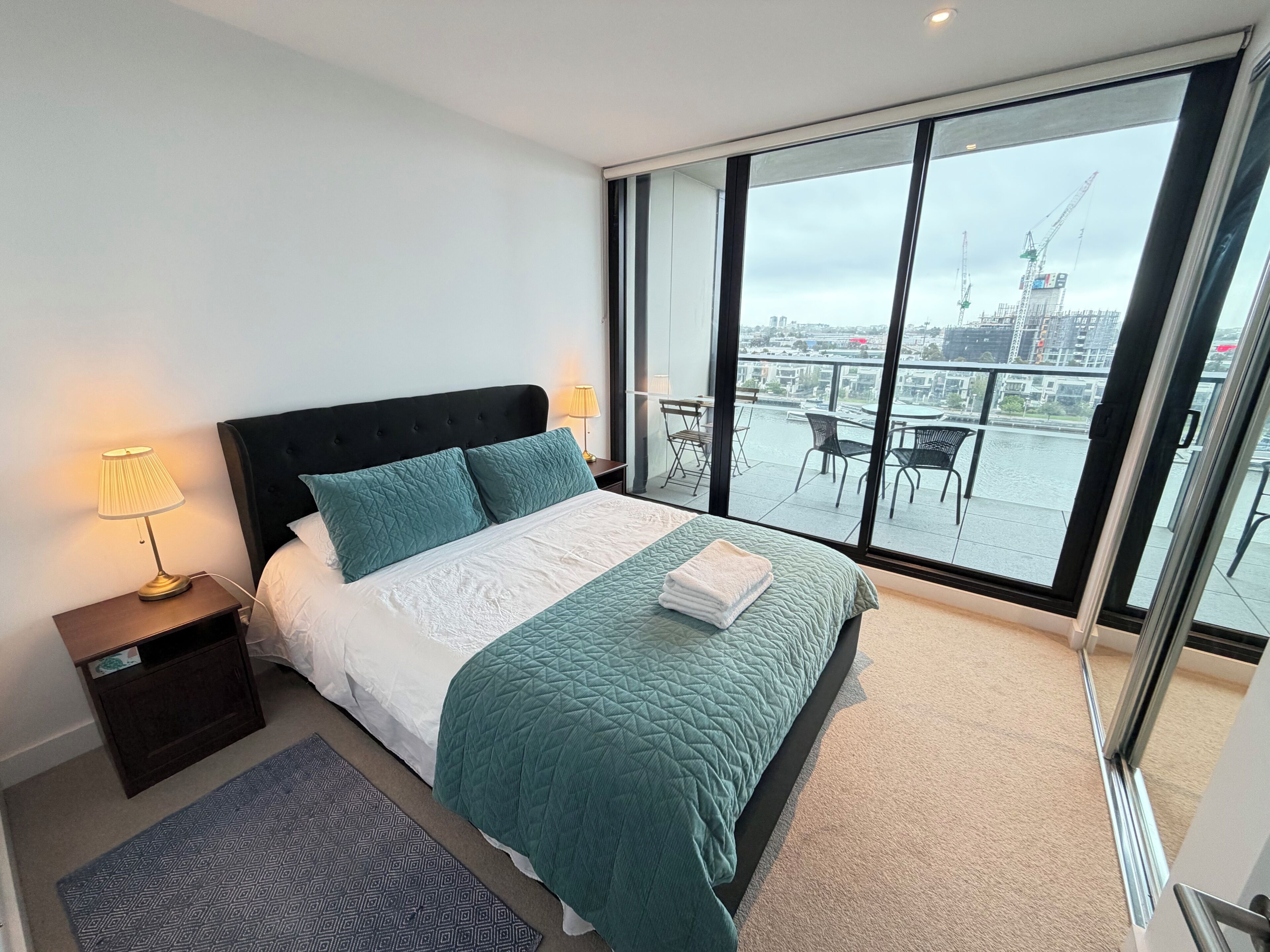A bedroom with a large bed, a nightstand, a lamp, a patterned rug, and a view of a balcony with outdoor furniture.