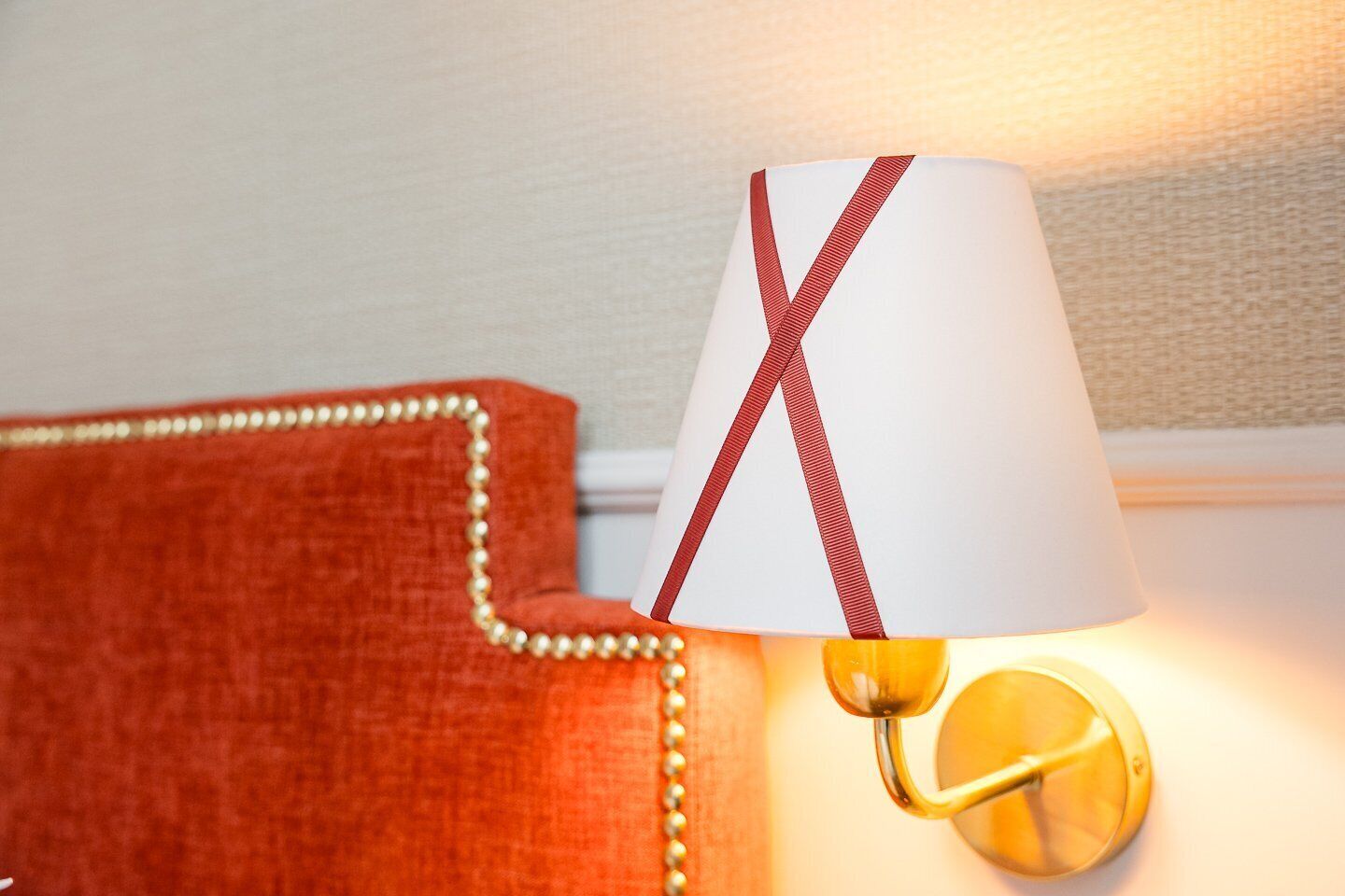 Une lampe avec un abat-jour blanc et un ruban rouge, placée à côté d’un fauteuil rouge recouvert de tissu, orné de galons dorés.
