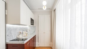 Una cocina estrecha con encimera de mármol, hervidor de agua y cafetera. Se observa un armario de madera y una puerta blanca al fondo.