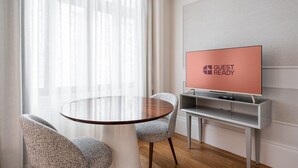 Una habitación moderna con una mesa redonda de vidrio, dos sillones tapizados, un televisor de pantalla plana sobre un mueble bajo y un ventanal con cortinas ligeras.