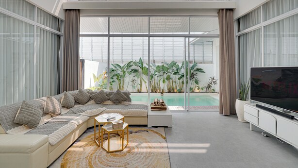 A modern living room with a sectional sofa, a flat-screen TV, and a large window overlooking a pool. -  Villa Osmanthus - Kerobokan (Kerobokan)