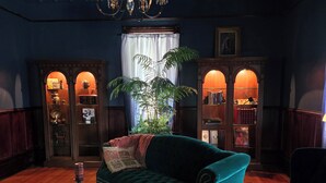 A cozy room with a green velvet sofa, a wooden coffee table, and a large plant. There are two wooden cabinets with books and decorative items. - Pinwheel Place off the Square (Talladega)