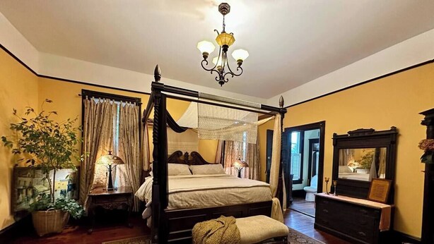 A four-poster bed with a canopy, a chandelier, and a large ornate rug in a room with wooden flooring and yellow walls. - Pinwheel Place off the Square (Talladega)