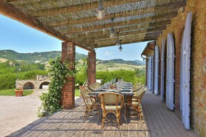 Un espace de restauration en plein air avec une table dressée pour six personnes, entourée de chaises en osier, sous une pergola offrant une vue sur des collines verdoyantes.