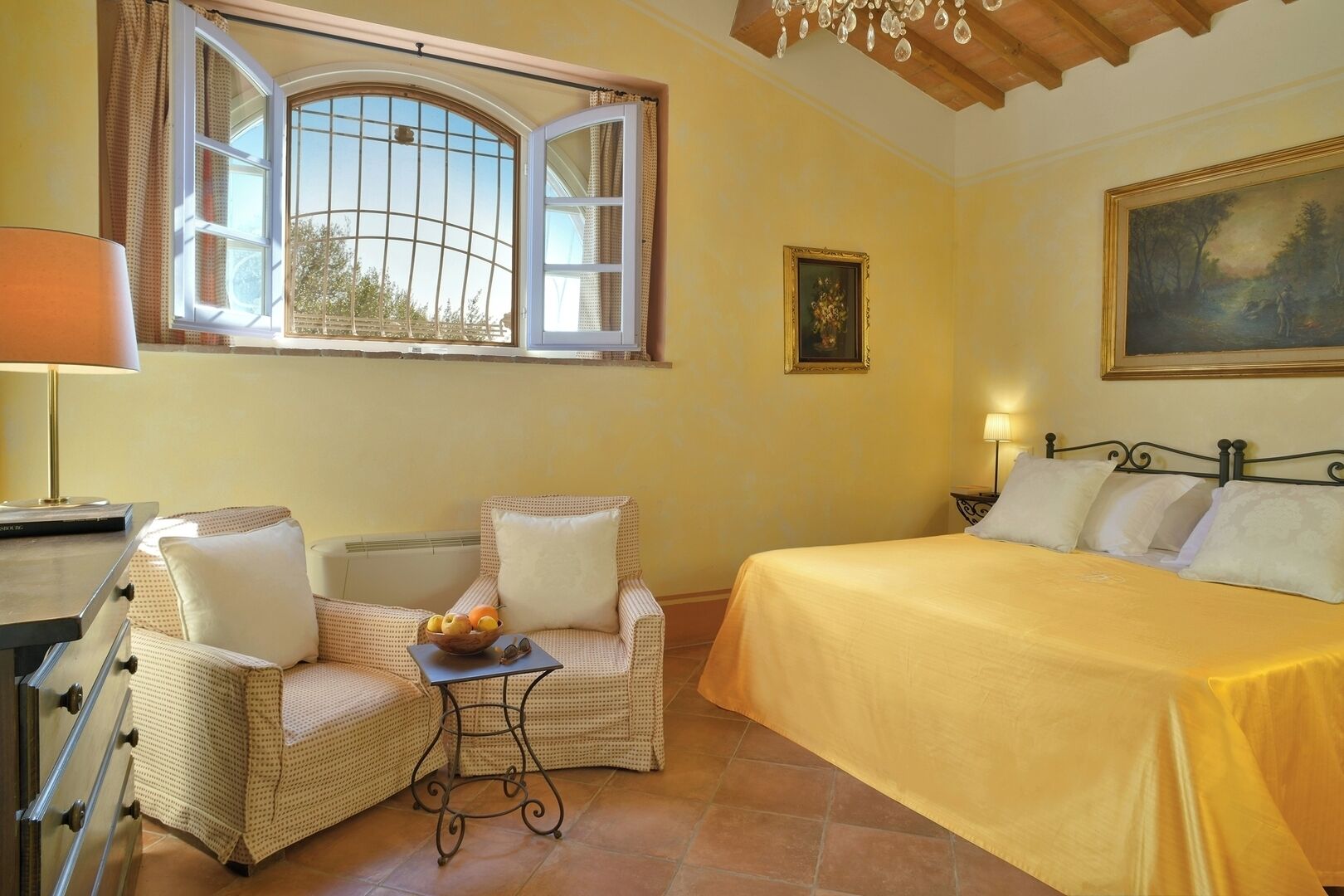 A bedroom with a bed, two chairs, a small table with fruit, a lamp, a painting, and a window with curtains.