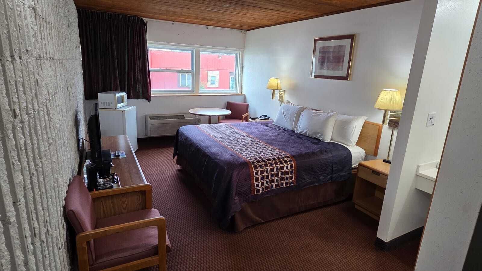 A hotel room with a bed, a desk, a chair, a table, two bedside lamps, and a window with a view of a red building.