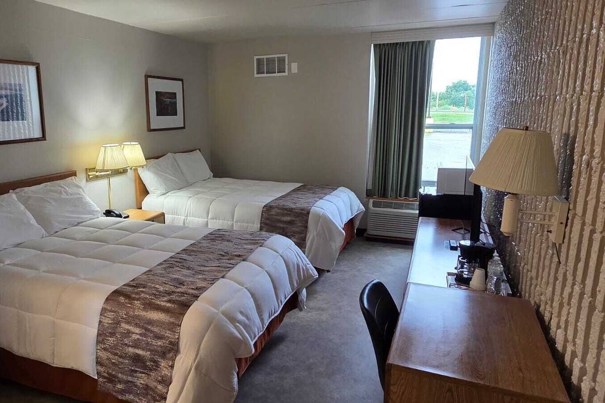 A hotel room with two beds, a desk, and a chair. There are two lamps on the desk and a window with curtains.
