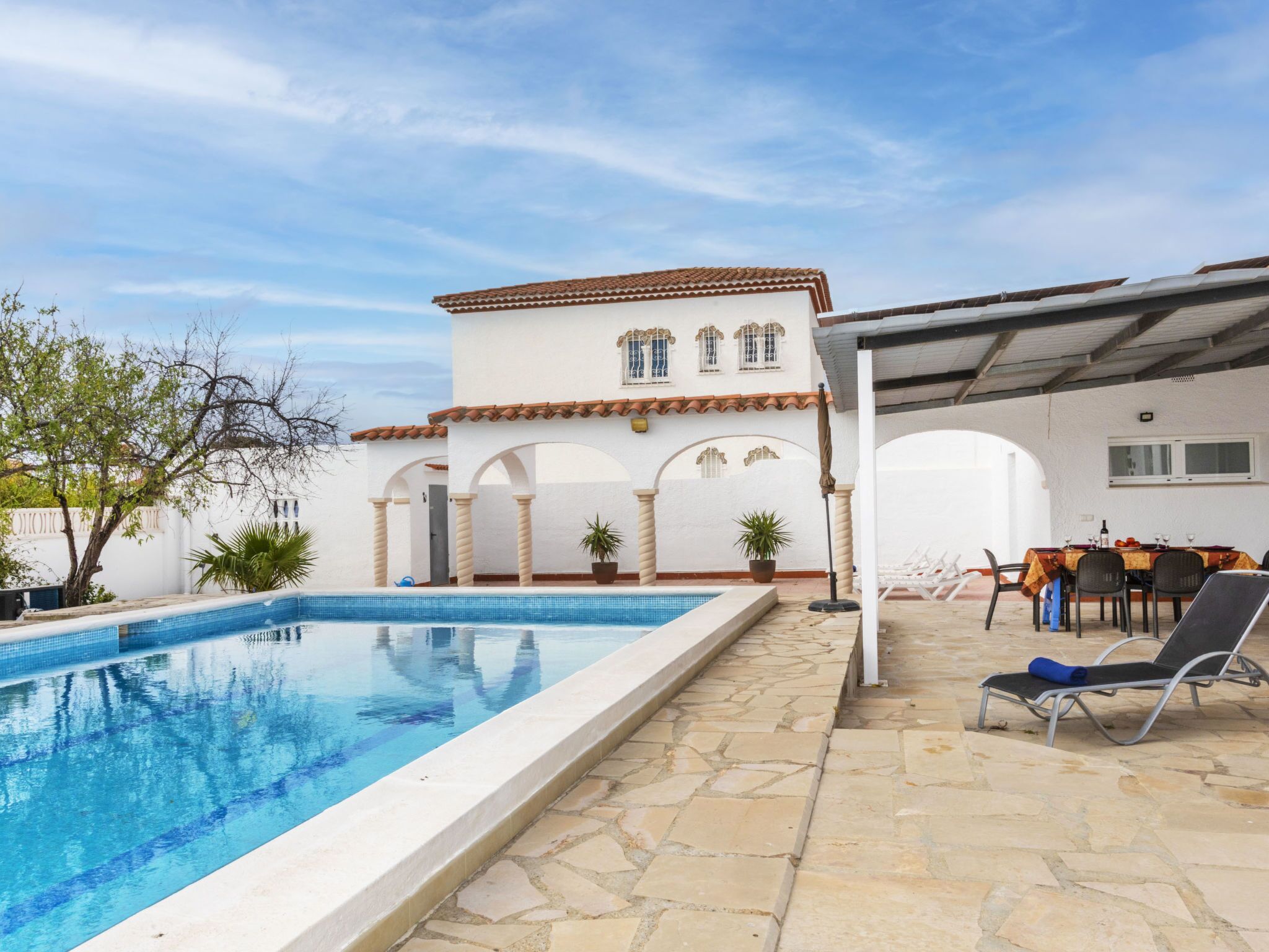 Una piscina con borde de baldosas, un porche cubierto con mobiliario exterior y un edificio con arcos y tejado de baldosas.