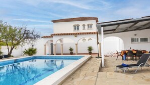 A swimming pool with a tiled deck, a covered patio with outdoor seating, and a building with arches and a tiled roof. - Junqué-Verdure by Interhome (Miami Platja)