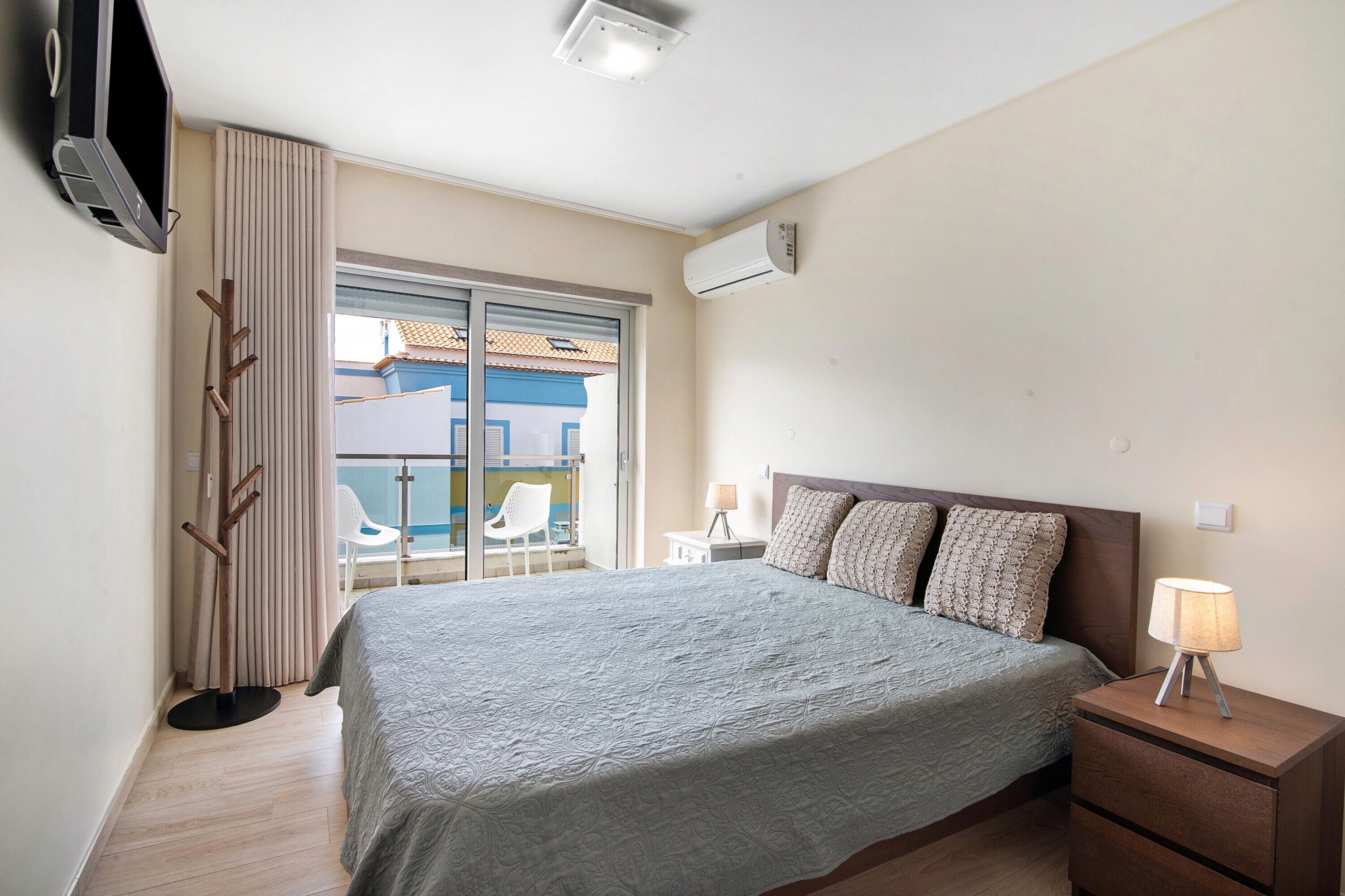 A bedroom with a bed, bedside table, lamp, air conditioning unit, TV, and a view of the outside.