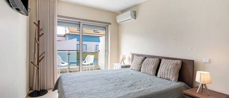 A bedroom with a bed, bedside table, lamp, air conditioning unit, TV, and a view of the outside.