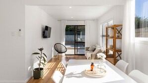 Una sala de estar moderna con un sofá blanco, una mesa de comedor con un centro de mesa y un amplio ventanal con persianas.