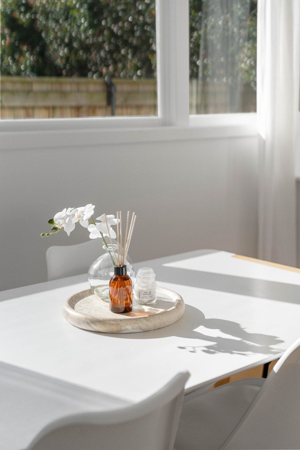 A table with a white tablecloth, a glass vase with white flowers, and a small bottle on a round tray.