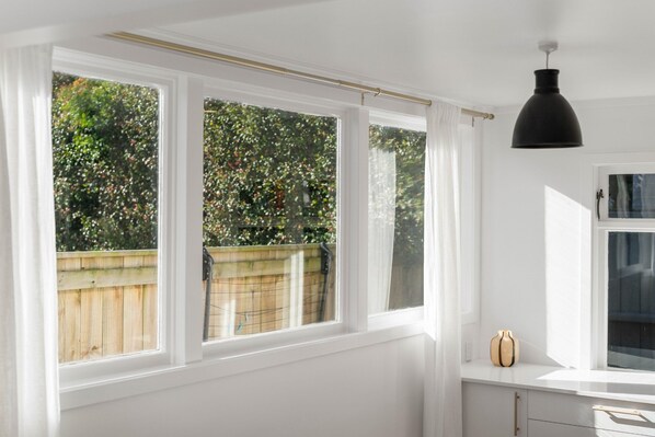 Ein Zimmer mit weißen Wänden, einem großen Fenster mit weißen Rahmen, einer schwarzen Pendelleuchte und einer kleinen Vase auf dem Fensterbrett.