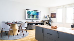 A modern living room with a round dining table, grey chairs, a flat-screen TV mounted on the wall, a wooden cabinet, and a small kitchen area with a microwave and toaster.